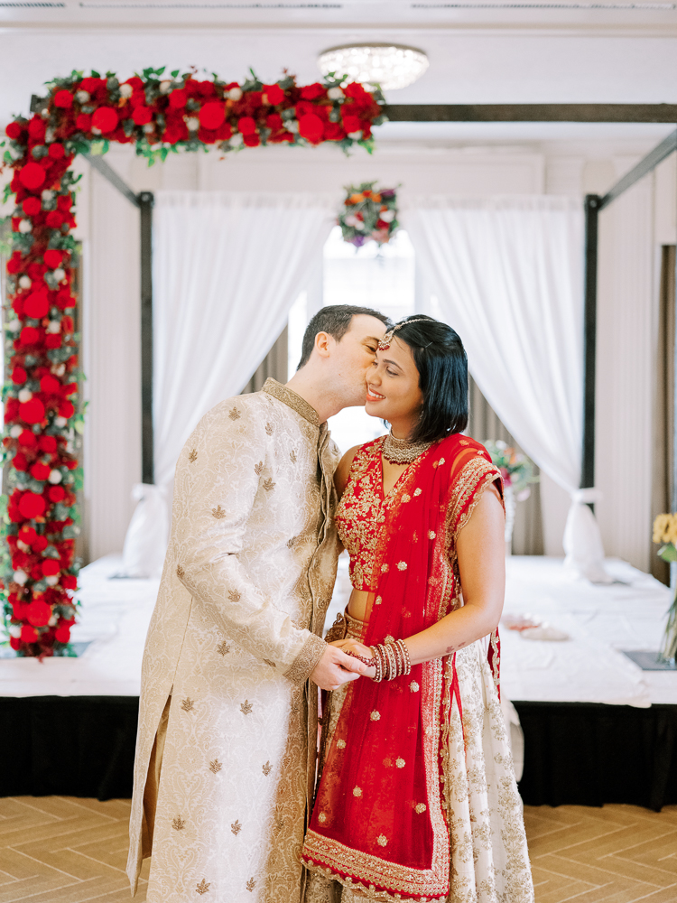 Indian Wedding Bride and Groom kiss by Hindu Ceremony with Red Florals in a Red and Tan Saree and Tan sherwani